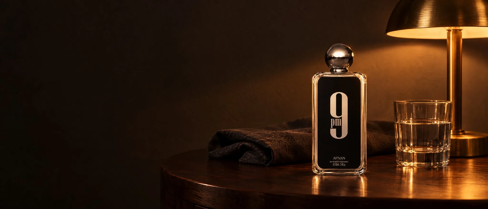 Afnan 9PM bottle on a dark wooden side table under a brass lamp, beside a glass of water